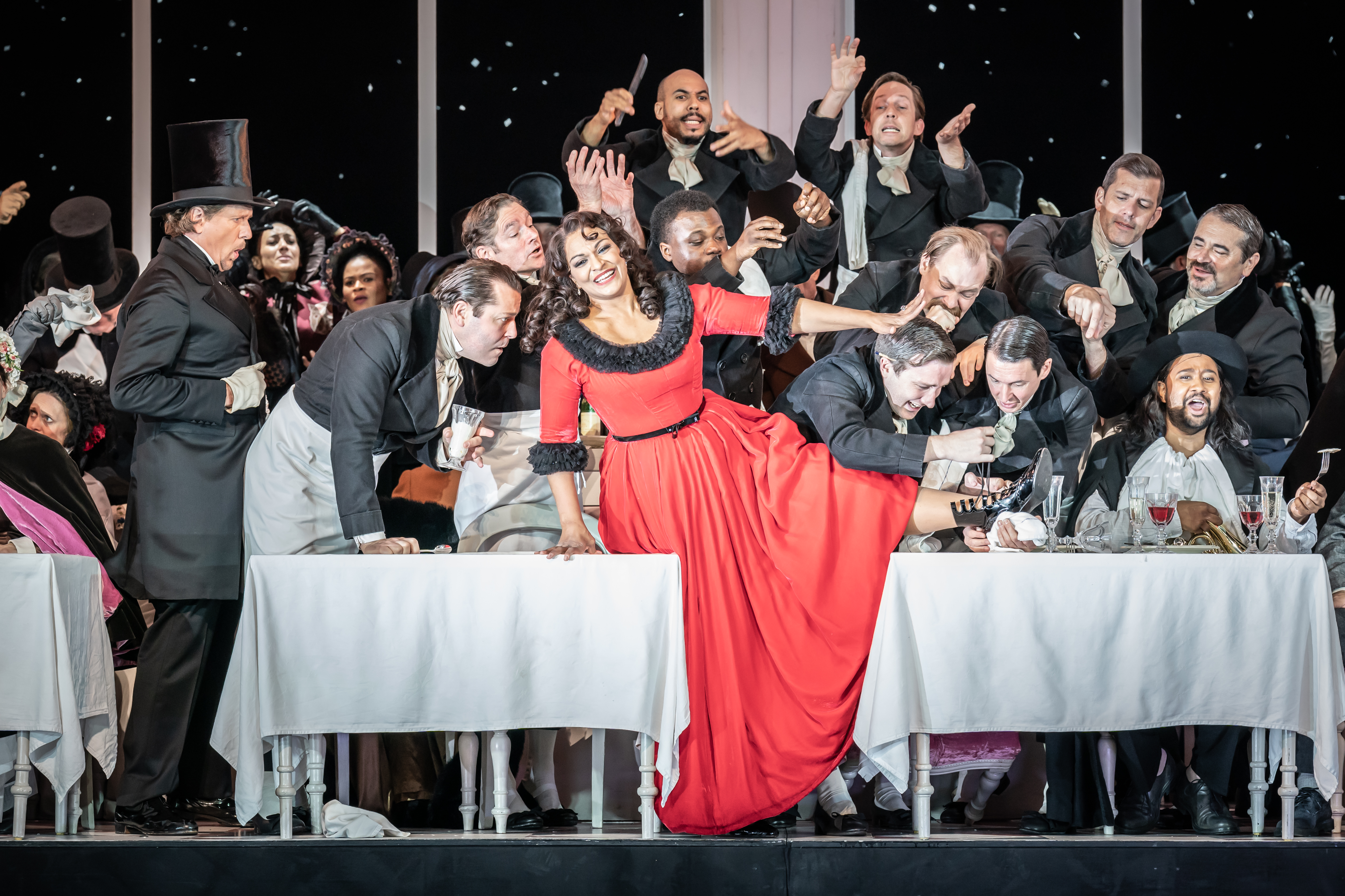 A woman in a red dress with black fur lining sits on a white draped table with several men around. 