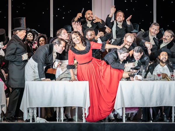 A woman in a red dress with black fur lining sits on a white draped table with several men around.