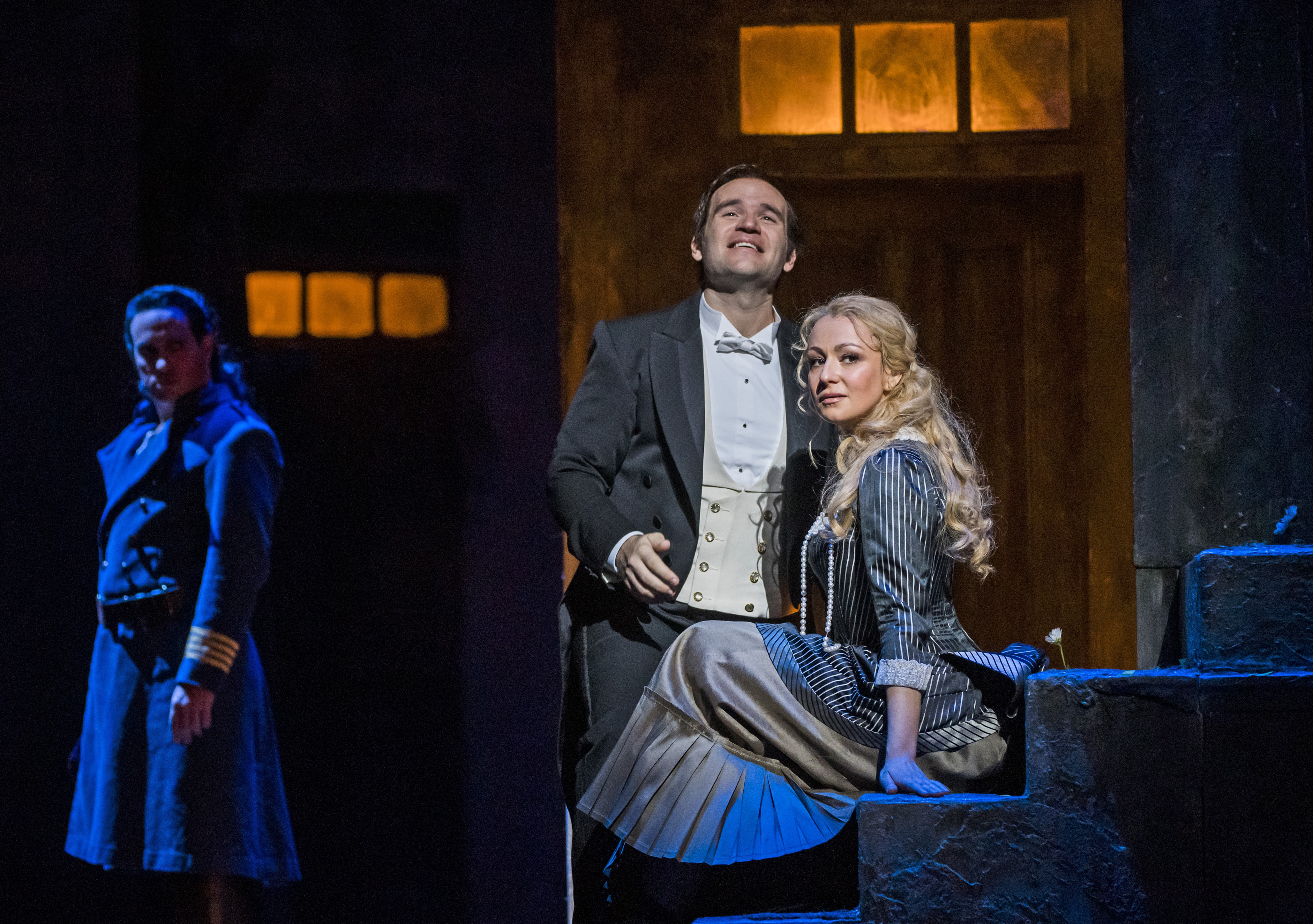 Irina Lungu performs as Marguerite and she sits on stone stairs wearing a pinstripe dress. Michael Fabiano performing as Faust stands closely by her side. He wears a suit and bow tie and he is gazing upwards. The pair are illuminated by a spotlight, while in the shadows Erwin Schrott as Méphistophélès watches on.