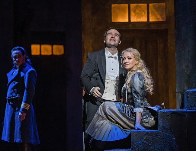 Irina Lungu performs as Marguerite and she sits on stone stairs wearing a pinstripe dress. Michael Fabiano performing as Faust stands closely by her side. He wears a suit and bow tie and he is gazing upwards. The pair are illuminated by a spotlight, while in the shadows Erwin Schrott as Méphistophélès watches on.