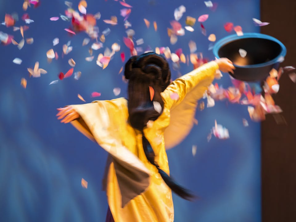 A woman in a traditional Japanese Geisha costume has her arms outstretched as she spins. She is holding a black bowl, and pink cherry blossom petals are falling around her as she turns.
