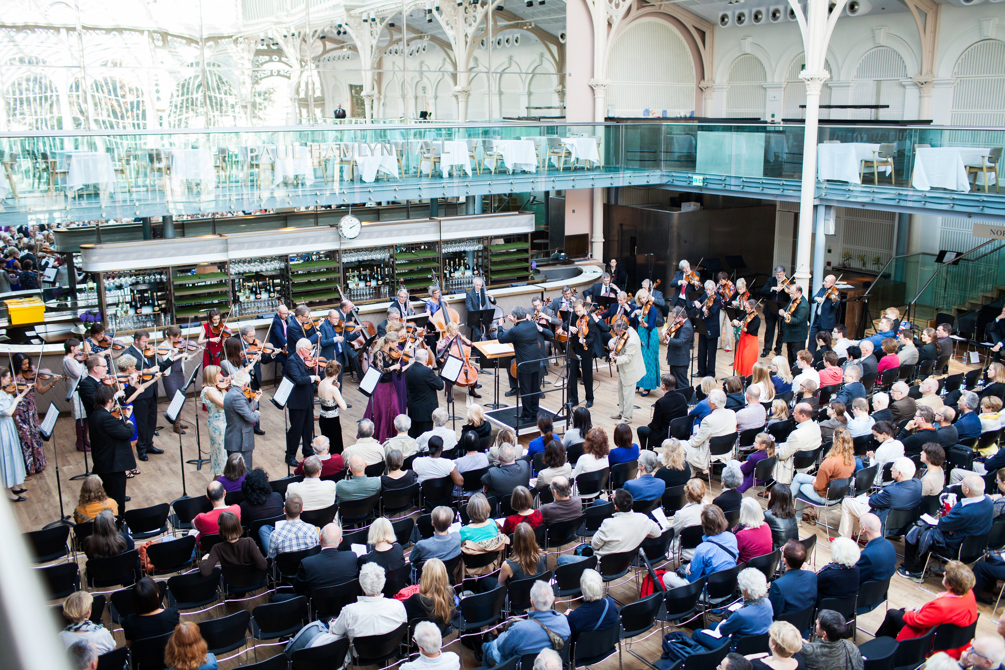 A crowd of people sit to watch a small orchestra perform inside a glass covered room which is the Paul Hamlyn Hall of the  Royal Opera House.