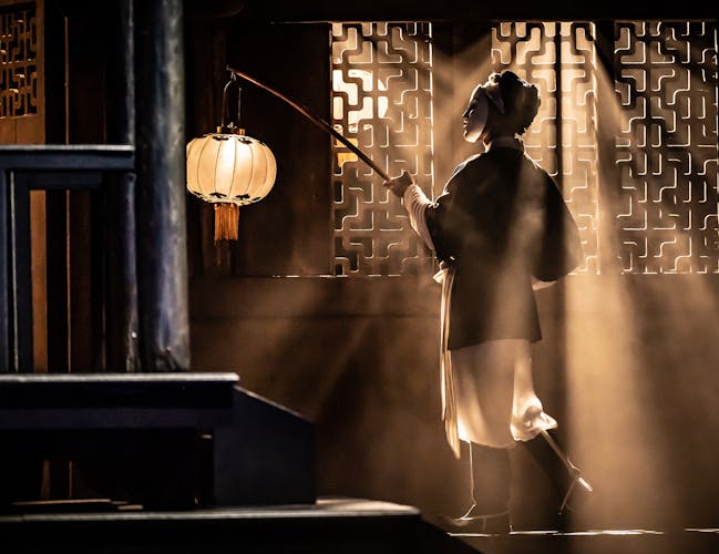 A character in Oriental costume holds a lit lantern with their back to the audience, light flows through a window