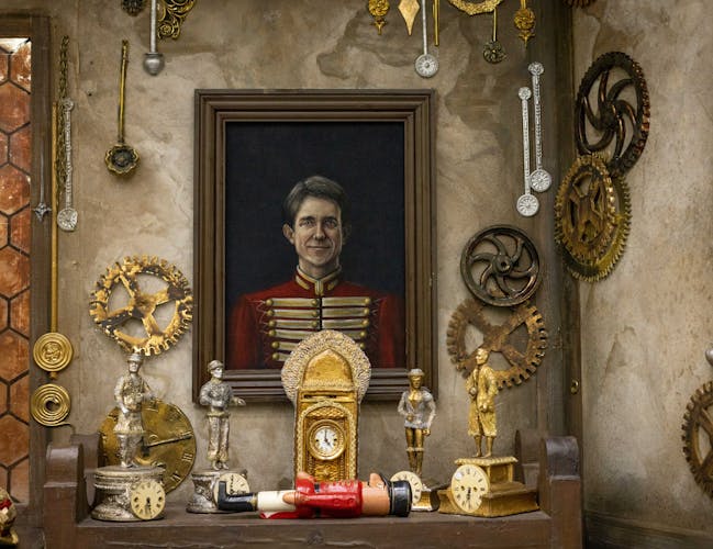 A collection of props taken backstage of the Royal Ballet production of The Nutcracker. A painted portrait of a smiling man in a red military uniform hangs on stained brown wall. Metallic cogs hang all around the walls, and bronze statues sit on a table in front of the portrait. A wooden nutcracker doll lies in the centre on its back.
