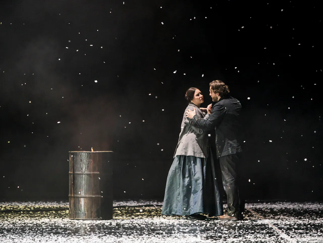 Two performers romantically embrace. One has long brown hair and is wearing a red dress with black fur lining and she holds onto the head of the other performer who is wearing a paint-specked artist smock jacket. They are Aida Garifullina performing as Musetta and Andrzej Filończyk as Marcello in The Royal Opera’s La bohème.