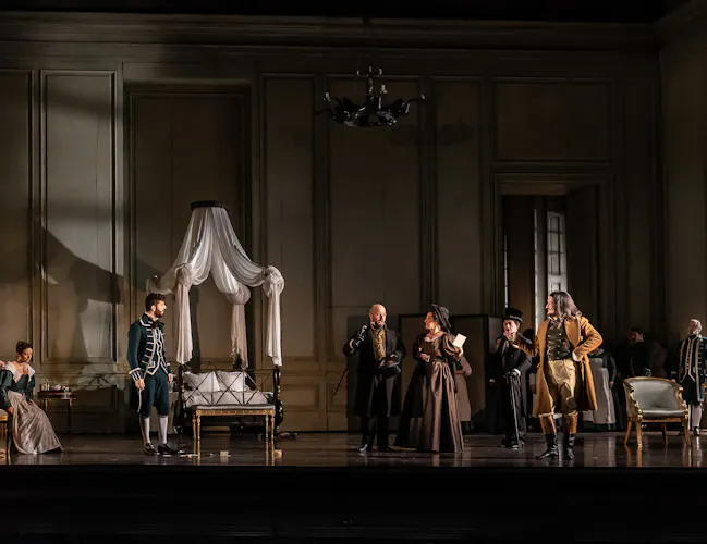 A group of people stand in a bedroom. All of them are looking towards a woman in a brown dress who is holding a letter.