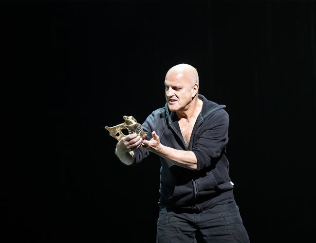 Actor Christopher Purves stands holding the lower portion of a skull in his hands and gazes upon it.