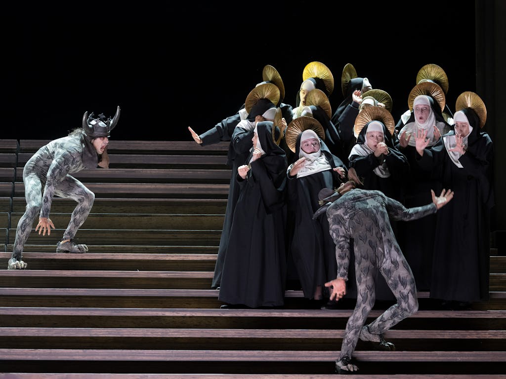 The Royal Opera performs Il Trovatore onstage. A group of characters dressed as nuns look on as two characters dressed in grey and wearing horns leap around them.