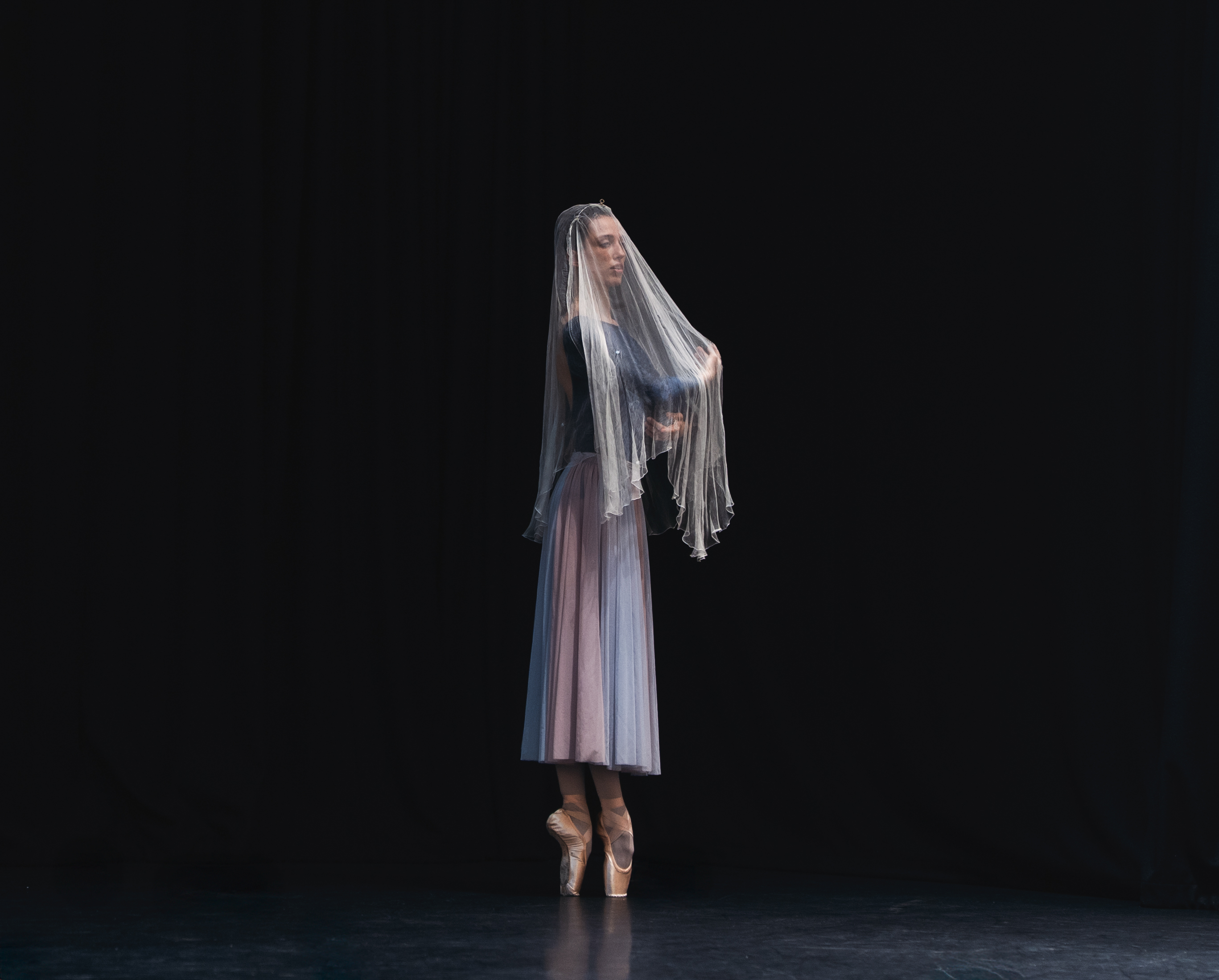 Nadia Mullova-Barley in rehearsal for Giselle. She stands en pointe and wears a blue leotard and white flowing skirt. She has a veil over her face.