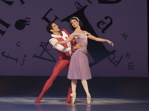 The ballet dancer Lauren Cuthbertson stands on stage en pointe wearing a purple dress, behind her with their heads around her waist is the ballet dancer Federico Bonnelli wearing a red and white costume with a red heart on the chest and a white frilled collar. They are performing in The Royal Ballet's production of Alice's Adventures in Wonderland in 2017.