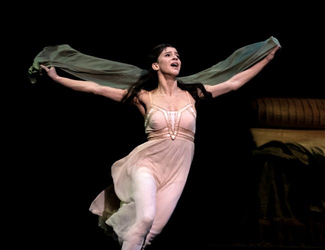 A ballet dancer in a pink dress with long black hair holds their arms upwards holding a shawl.