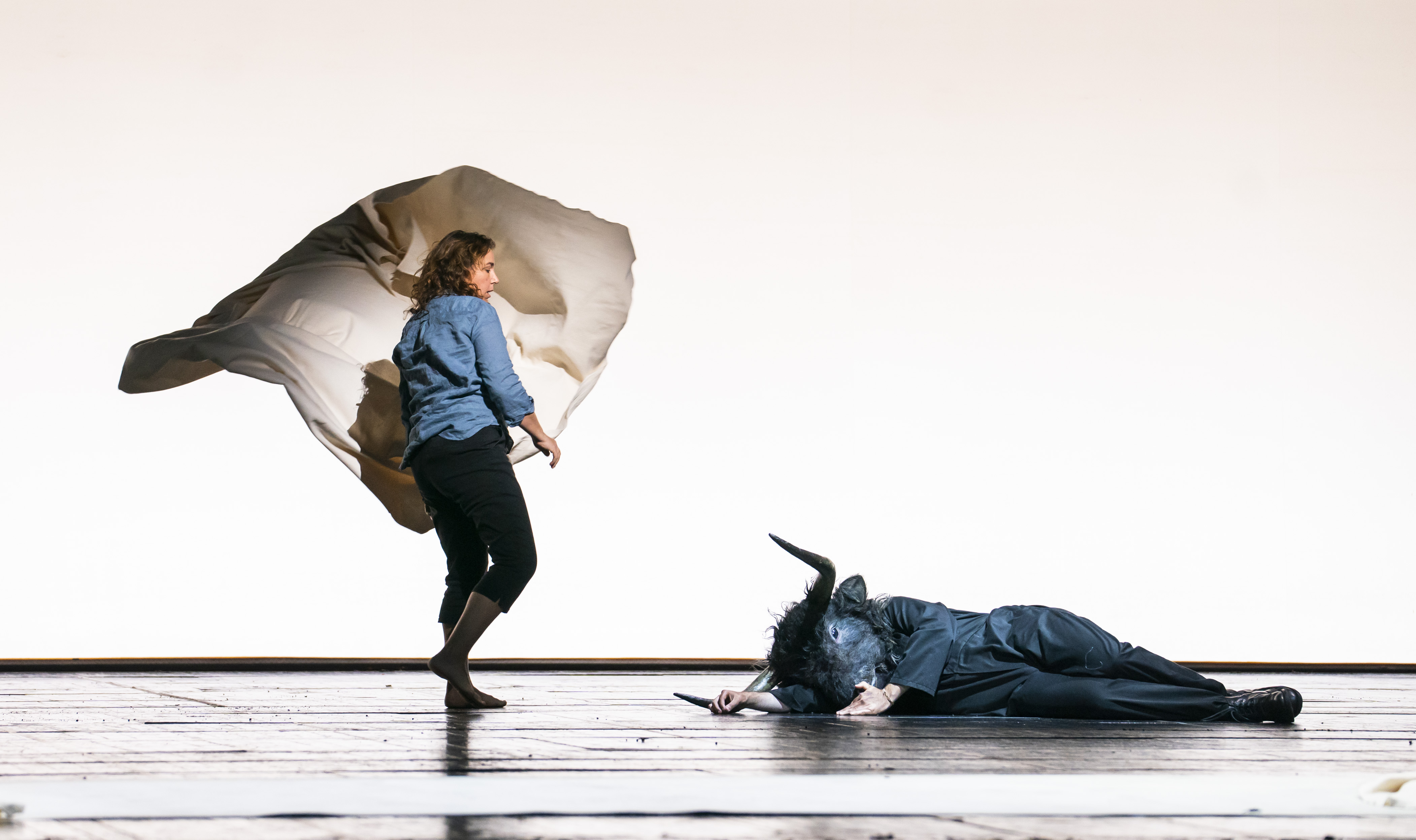 A production of the opera/ballet Phaedra and The Minotaur being performed on stage. A woman stands barefoot in jeans and a blue top, waving a white sheet. A monster with a body of a man and a head of a bull lies collapsed on the floor.