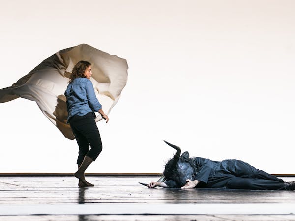 A production of the opera/ballet Phaedra and The Minotaur being performed on stage. A woman stands barefoot in jeans and a blue top, waving a white sheet. A monster with a body of a man and a head of a bull lies collapsed on the floor.