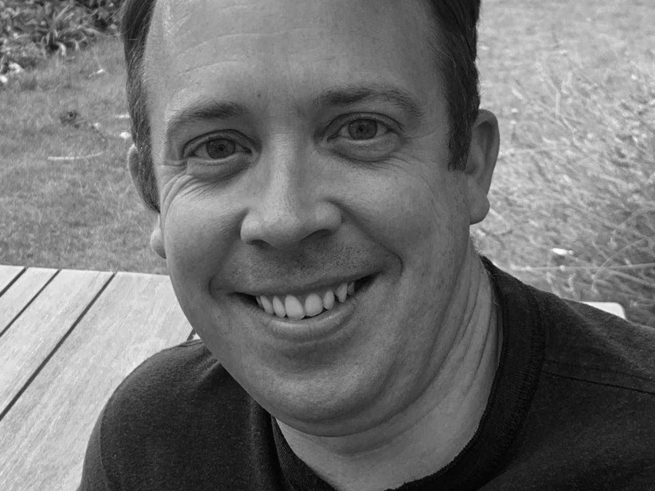 A black and white headshot of the illusions specialist Chris Fisher. He sits at a wooden table in a garden.