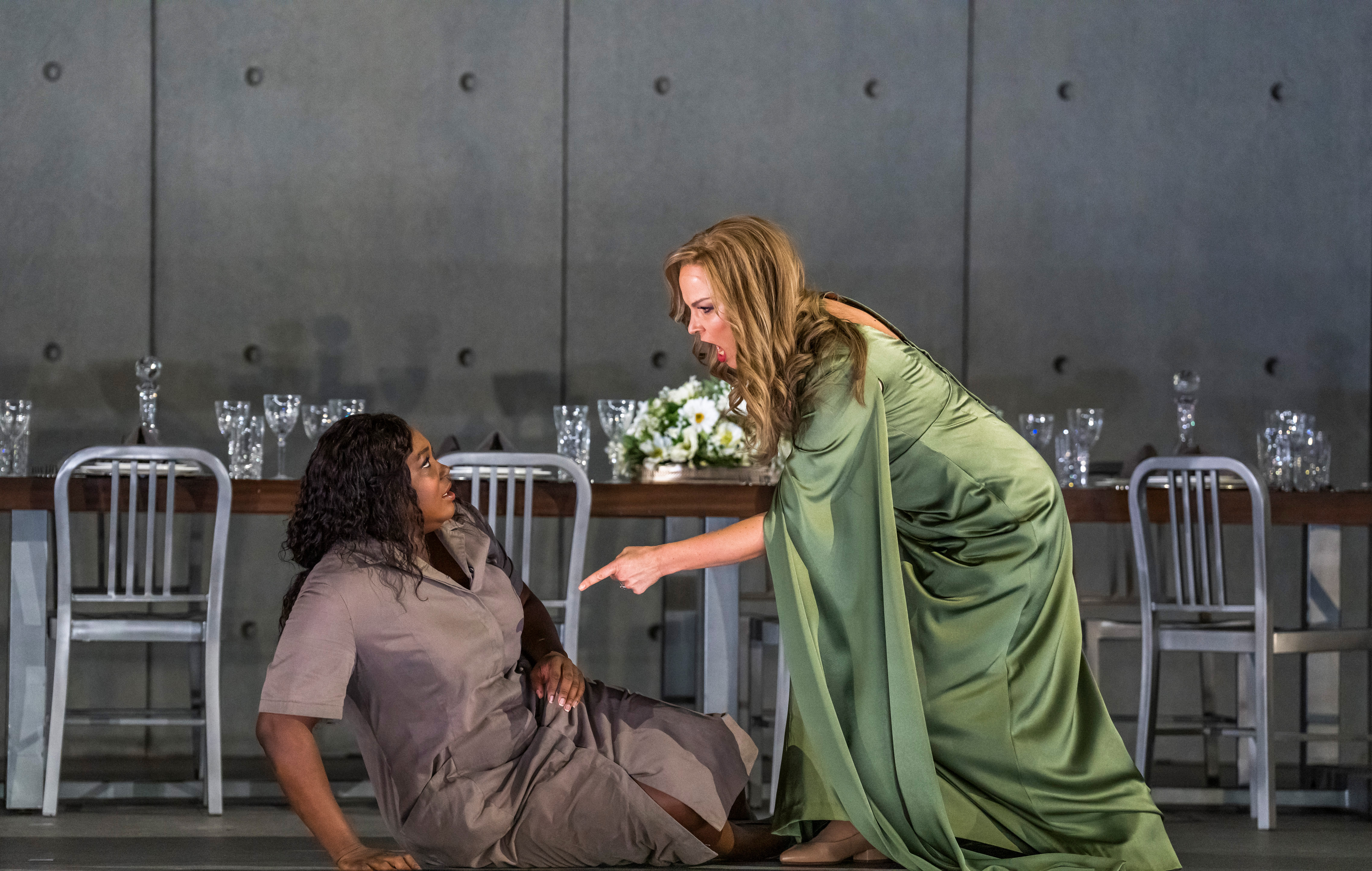 A production of the opera Aida is being performed onstage. A blonde woman in a flowing green dress stands menacingly, pointing a finger as she stands above a brunette dressed in a plain grey dress and sitting on the floor. 