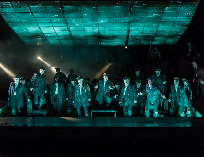 A group of sailors look out over the audience. They are bathed in green light as though light is reflecting off the water in front of them.