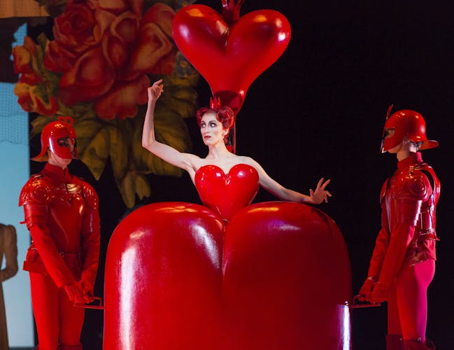 A production of Alice's Adventures in Wonderland performed by the Royal Ballet. A dancer as the Queen of Hearts gives a wide eyed stare to two other dancers dressed as her footmen. The costumes are a vivid red, and the Queen emerges from an enormous heart shaped dress.