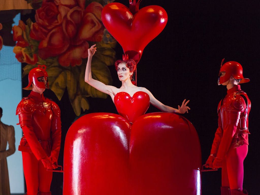 A production of Alice's Adventures in Wonderland performed by the Royal Ballet. A dancer as the Queen of Hearts gives a wide eyed stare to two other dancers dressed as her footmen. The costumes are a vivid red, and the Queen emerges from an enormous heart shaped dress.