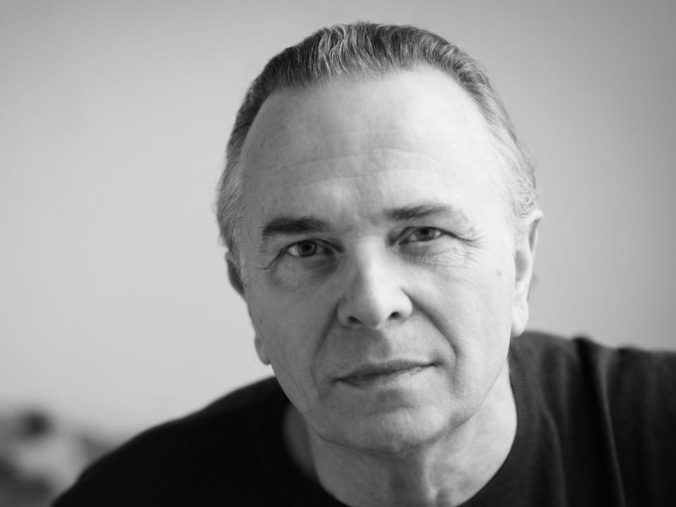 headshot of Sir Mark Elder in black and white. He's wearing a black tshirt and looking out at the camera.