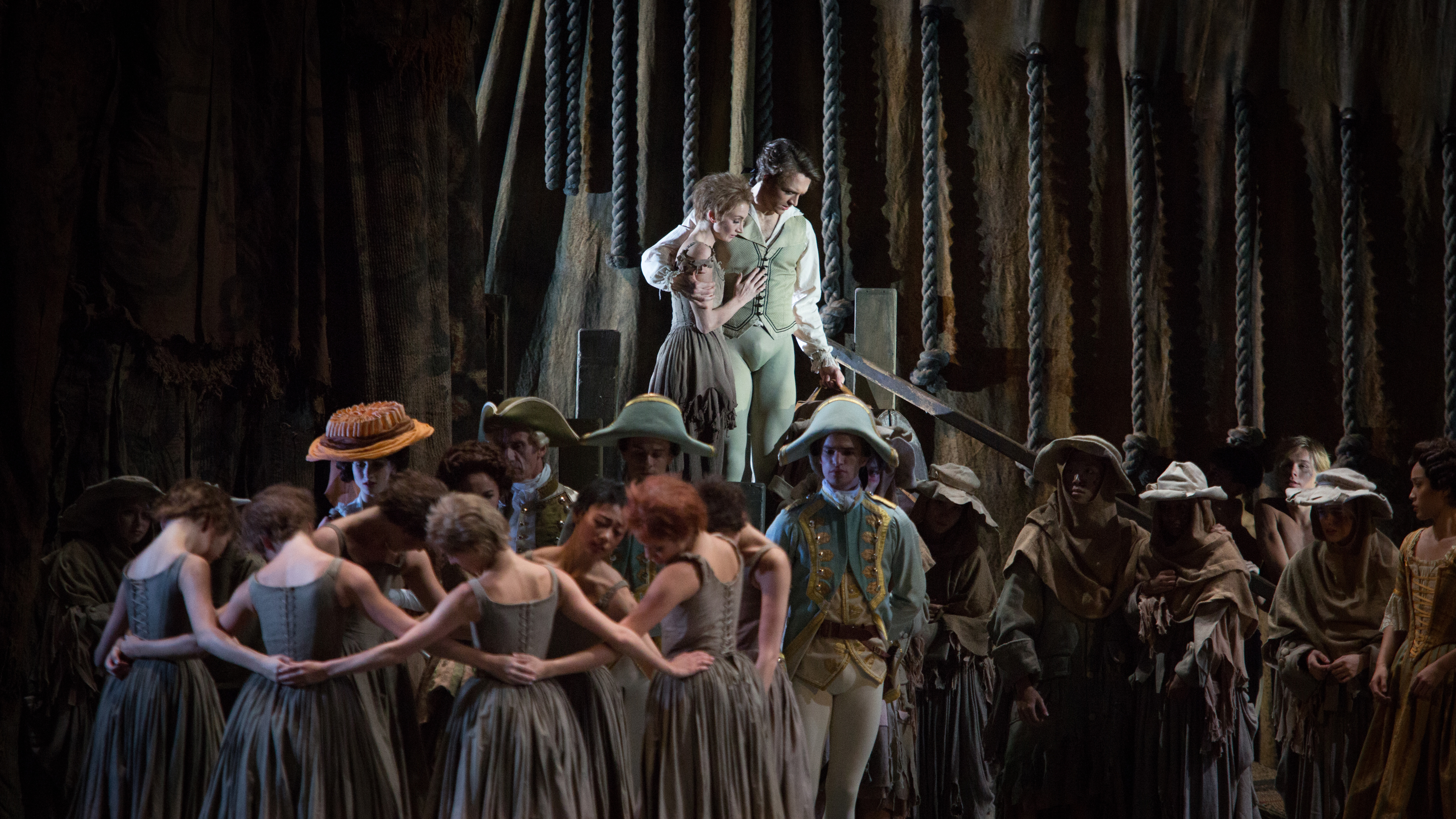 A group of Royal Ballet artists surround two dancers standing on a platform. 