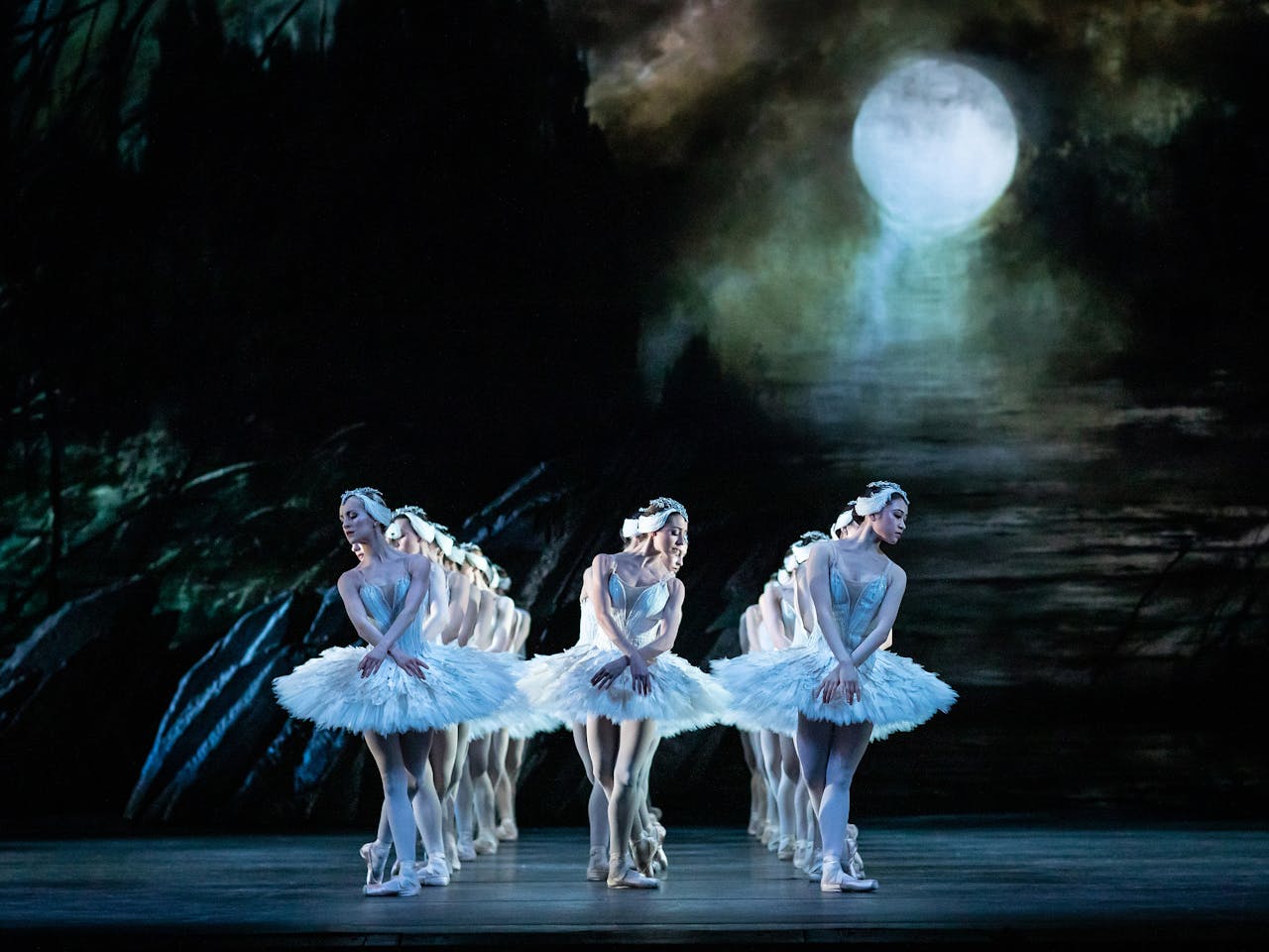 A group of Royal Ballet artists dance onstage in white tutus in a performance of Swan Lake. The dancers are dressed white feathers and matching headresses. The set has a glowing white moon and the dancers are arranged in three lines.