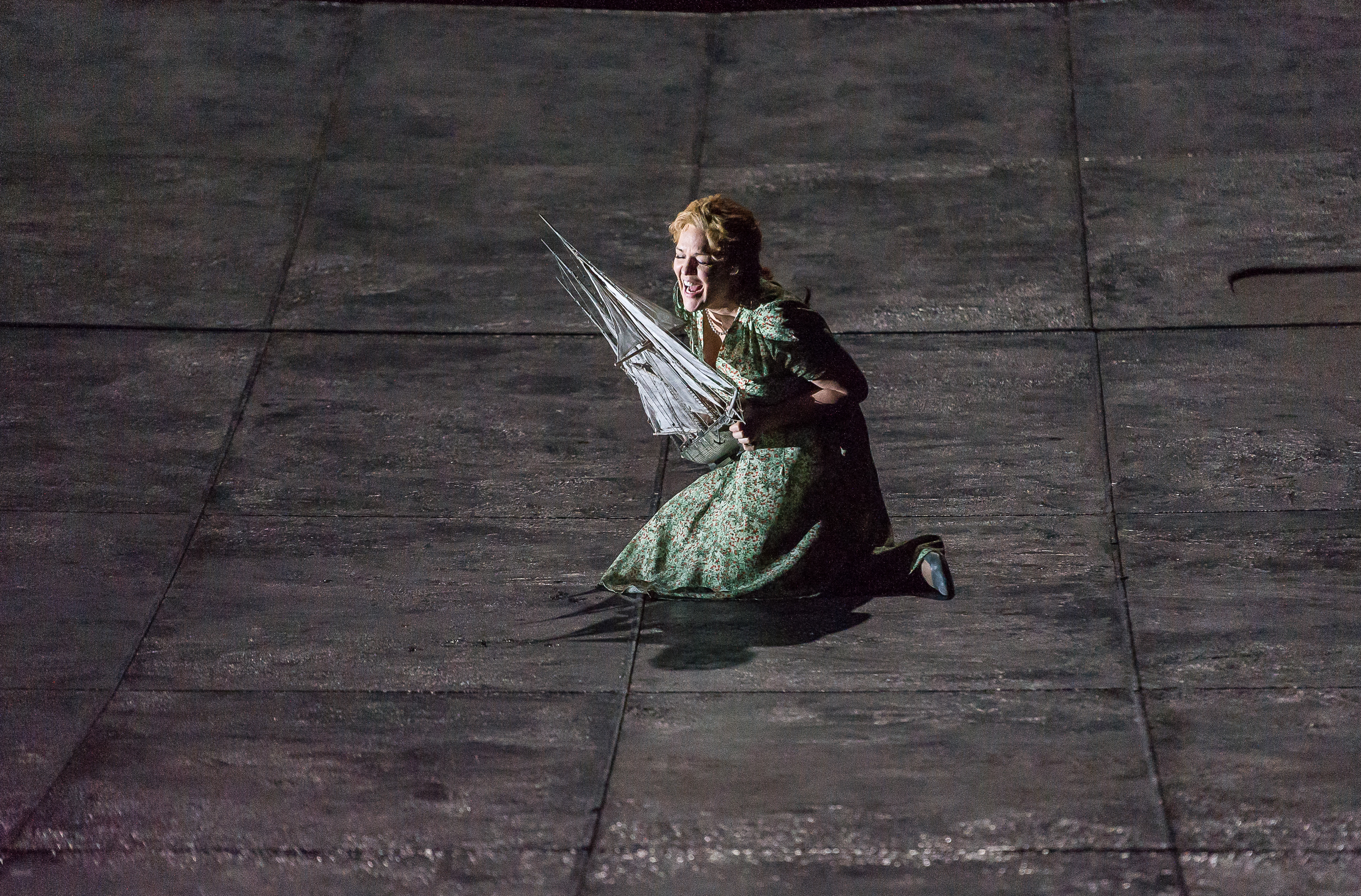 A woman kneels on a grey stage holding a model to her chest. 