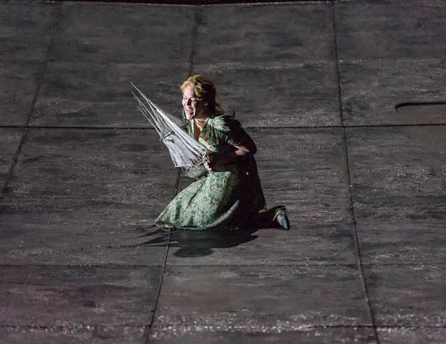 A woman kneels on a grey stage holding a model to her chest.