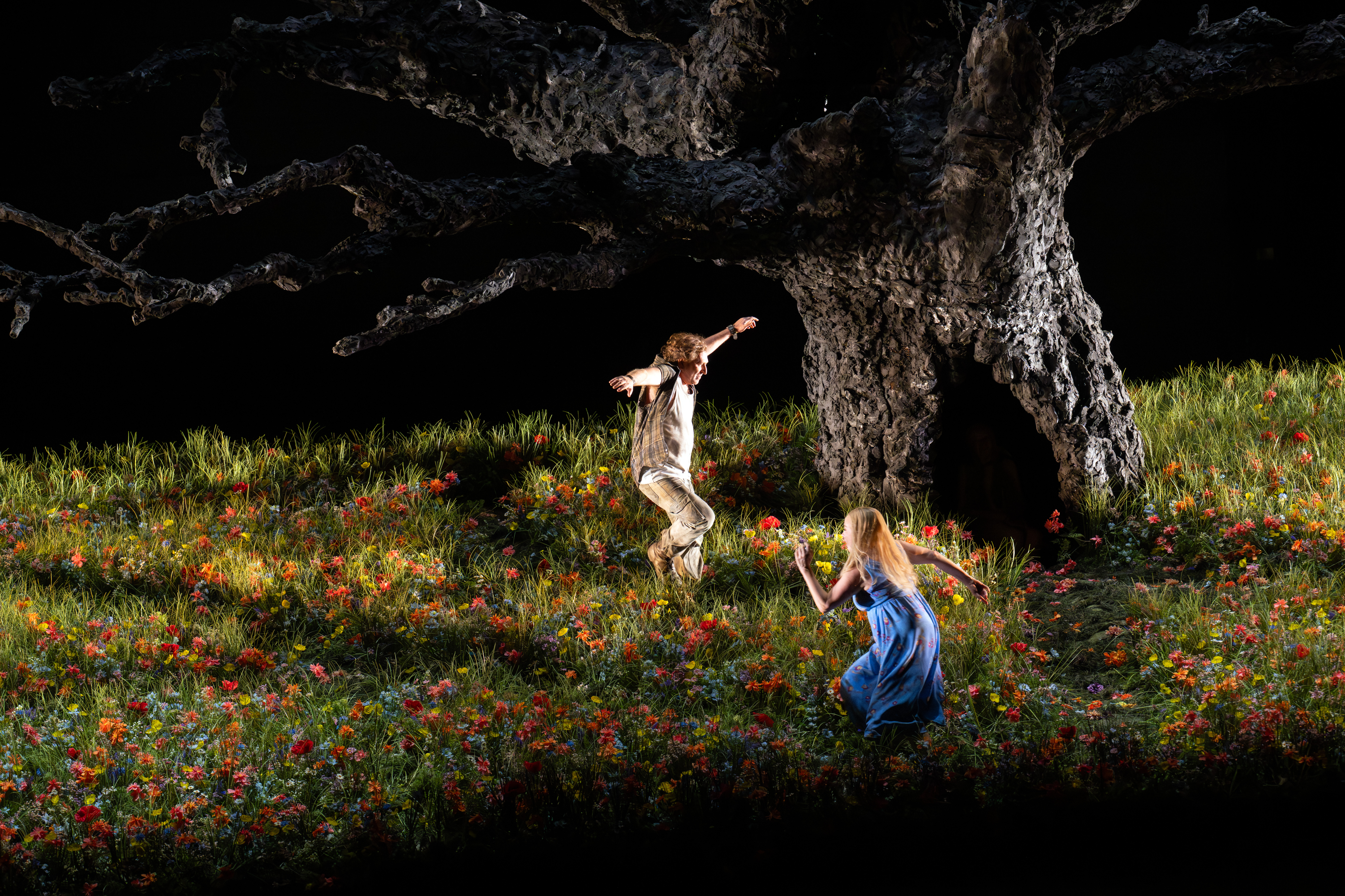 Two people run through a meadow on a dark stage. In the middle of the meadow is a large tree, it has no leaves. There is a large opening at the base of the tree.