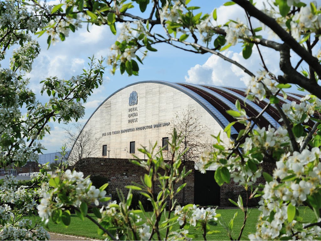 A building with a curved roof and the Royal Opera House logo on a beautiful day. The building is framed by white flowers as the camera focuses from across a bright green field.