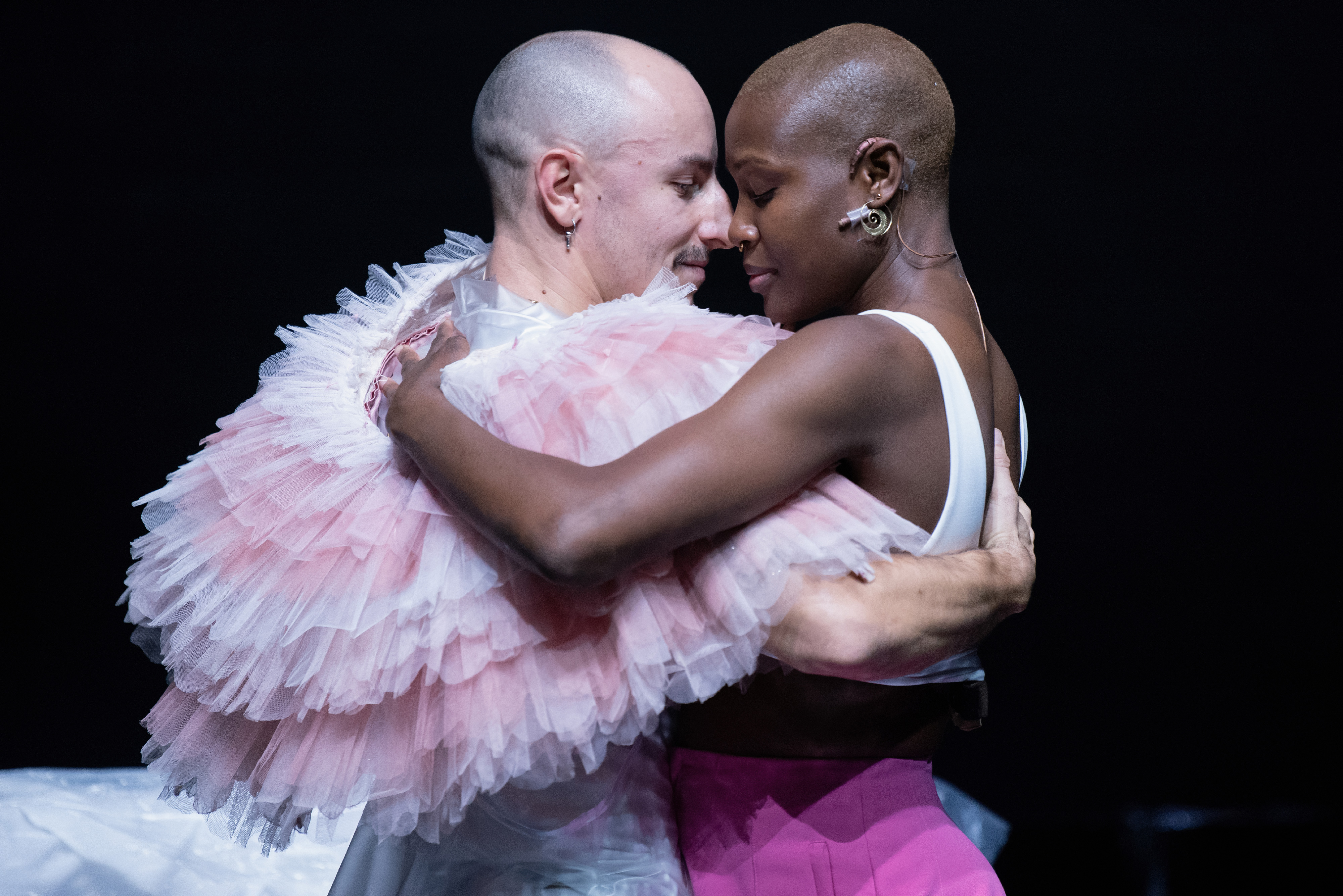 Two dancers embrace - one is wearing a frilly tutu around their neck, the other is wearing a white vest and pink skirt.