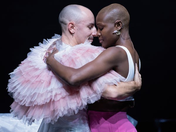 Two dancers embrace - one is wearing a frilly tutu around their neck, the other is wearing a white vest and pink skirt.