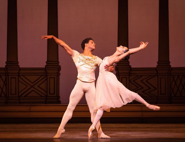 Two dancers in cream and gold costumes perform together, extending their arms and legs to the side