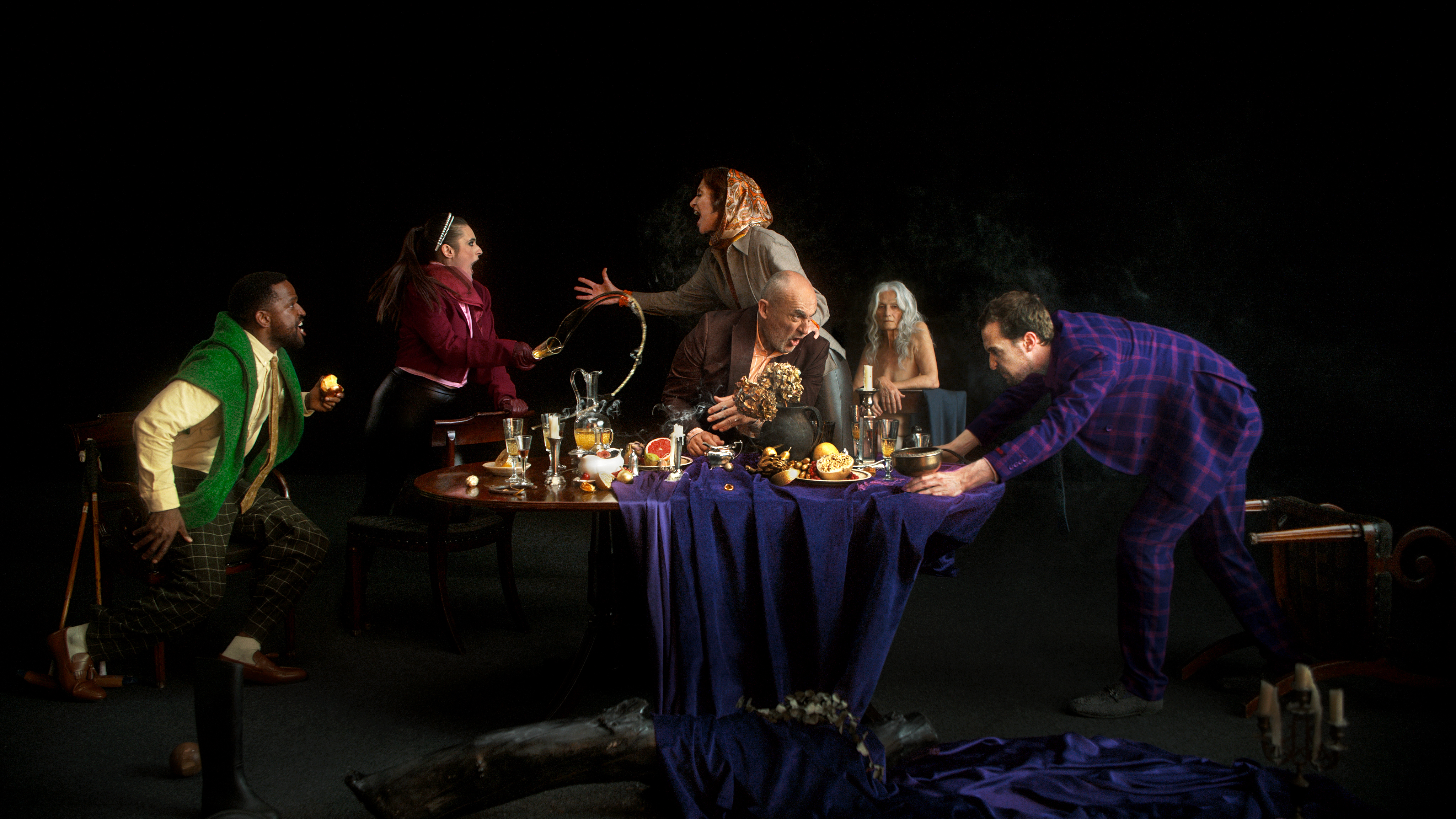 Several actors stand arguing around a grand dining table laid with food, drinks and flowers. One actor throws their drink while another pulls the table cloth from the table. In the background, an older lady sits observing the chaos.