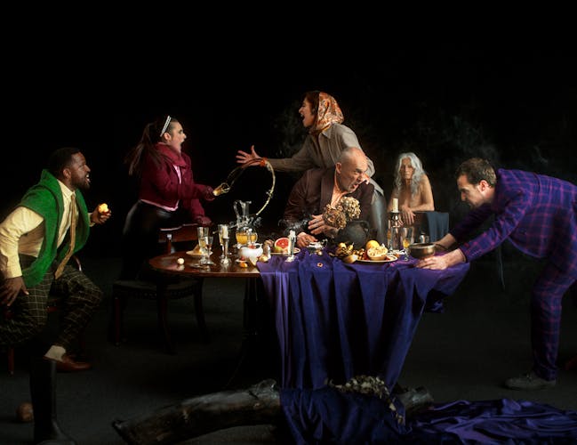 Several actors stand arguing around a grand dining table laid with food, drinks and flowers. One actor throws their drink while another pulls the table cloth from the table. In the background, an older lady sits observing the chaos.
