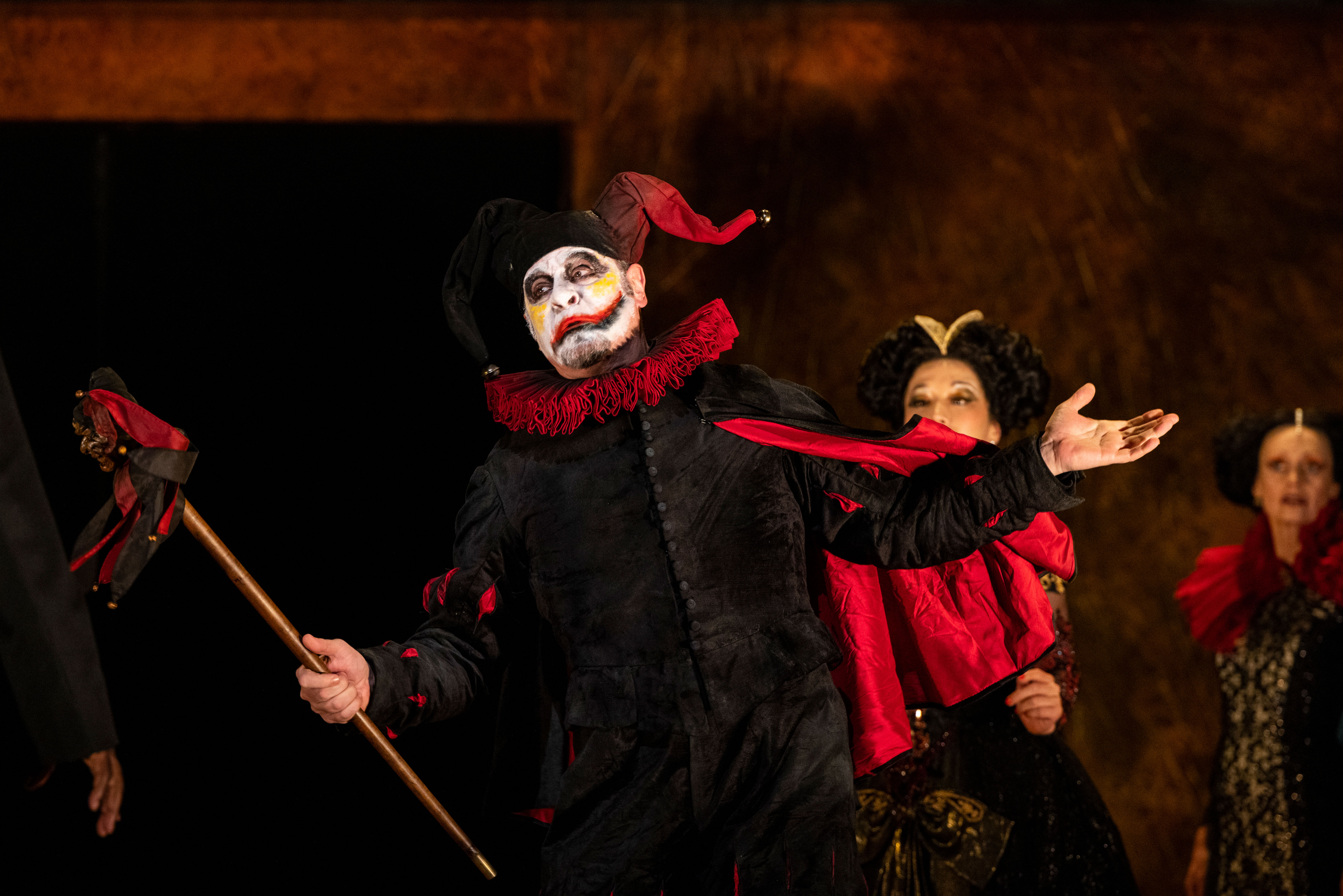 A clown in a black and red costume holds a staff. He has white face paint and a painted on grin. 