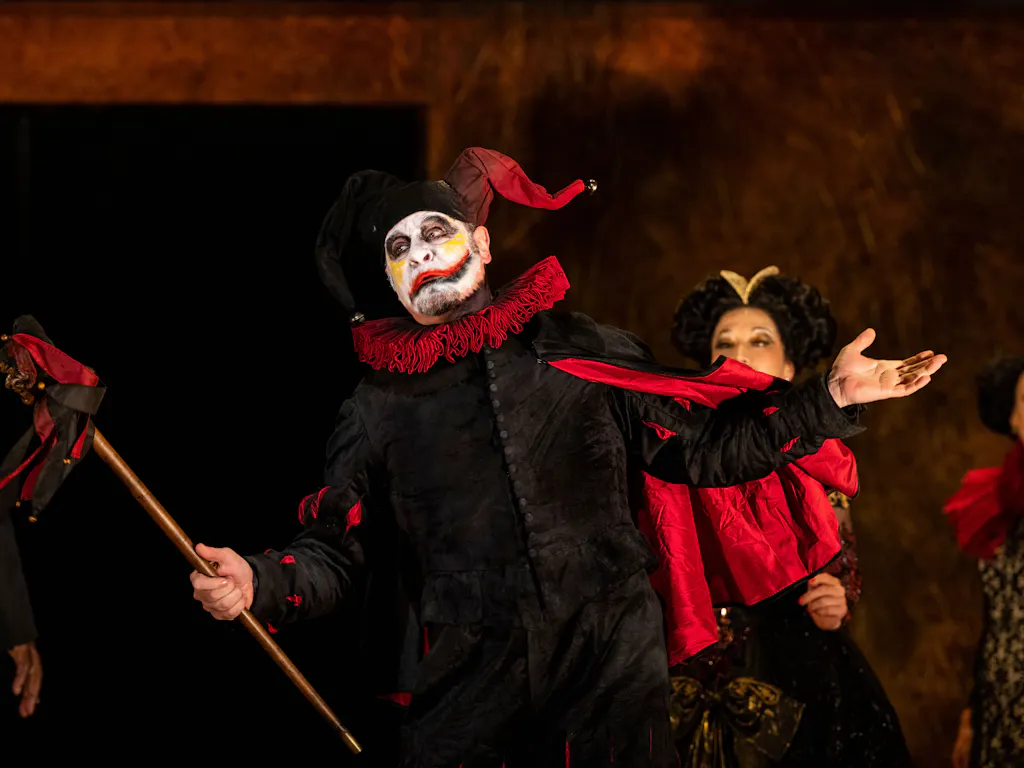 A clown in a black and red costume holds a staff. He has white face paint and a painted on grin.