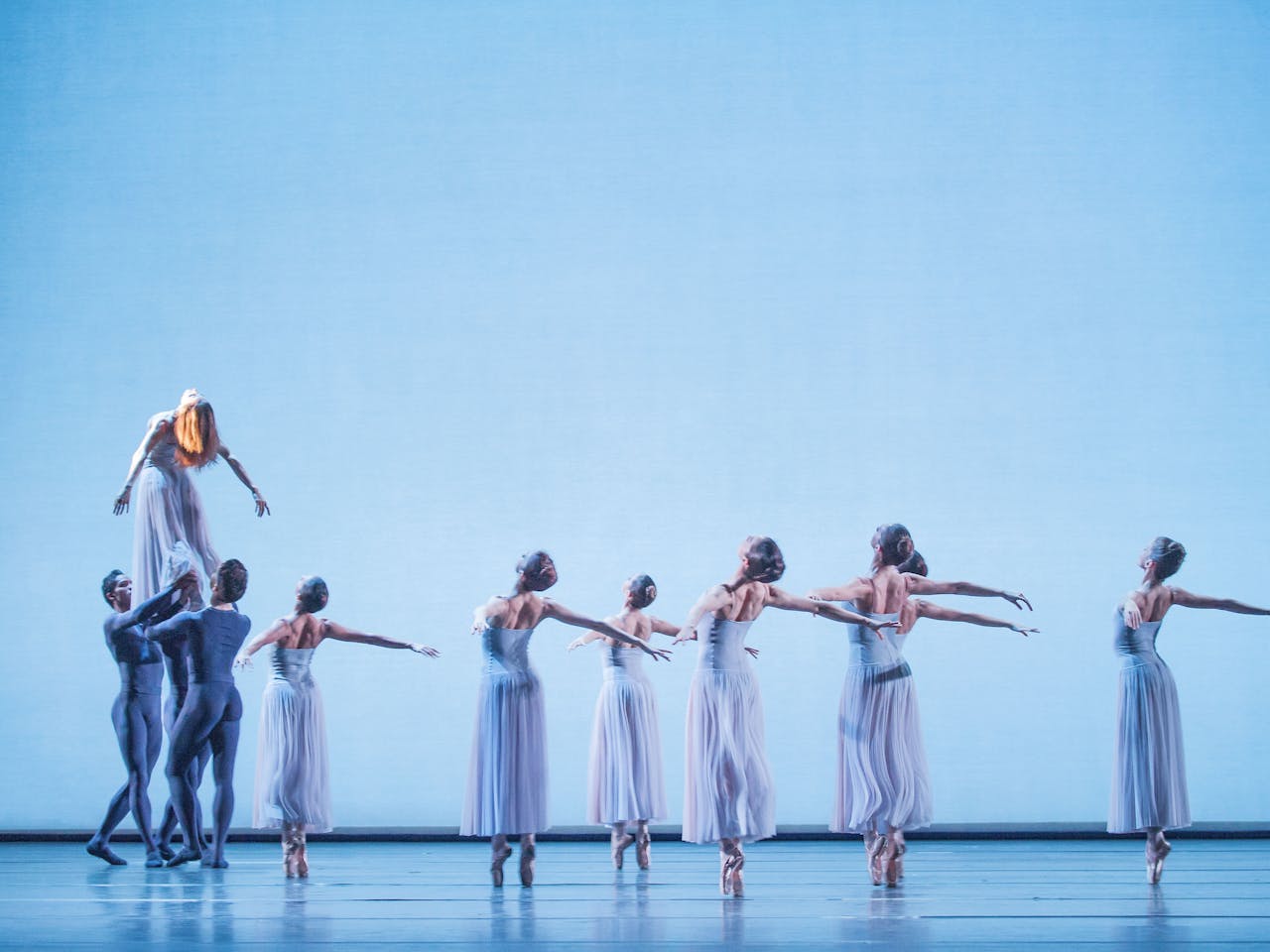 A group of ballerinas hold a pose on an empty stage. Their arms are outstretched behind them and they stand on their toes. On the left three male dancers hold a ballerina in the air.