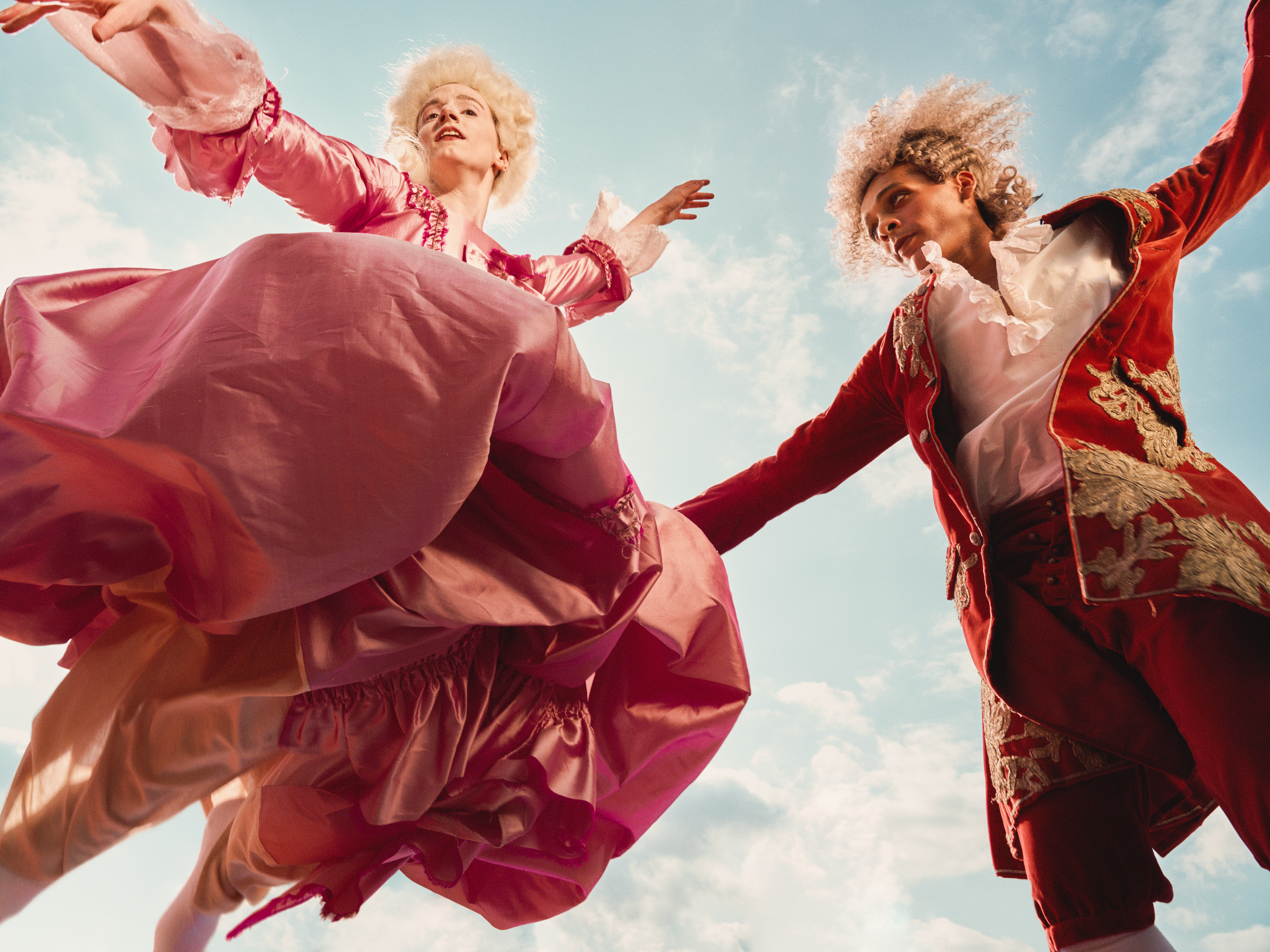 Two performers in elaborate pink and red period costumes leap above the camera against a bright blue sky, their arms outstretched and costumes billowing as they move. (2 of 4)