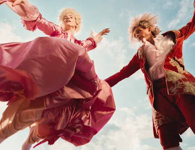 Two performers in elaborate pink and red period costumes leap above the camera against a bright blue sky, their arms outstretched and costumes billowing as they move. (2 of 4)