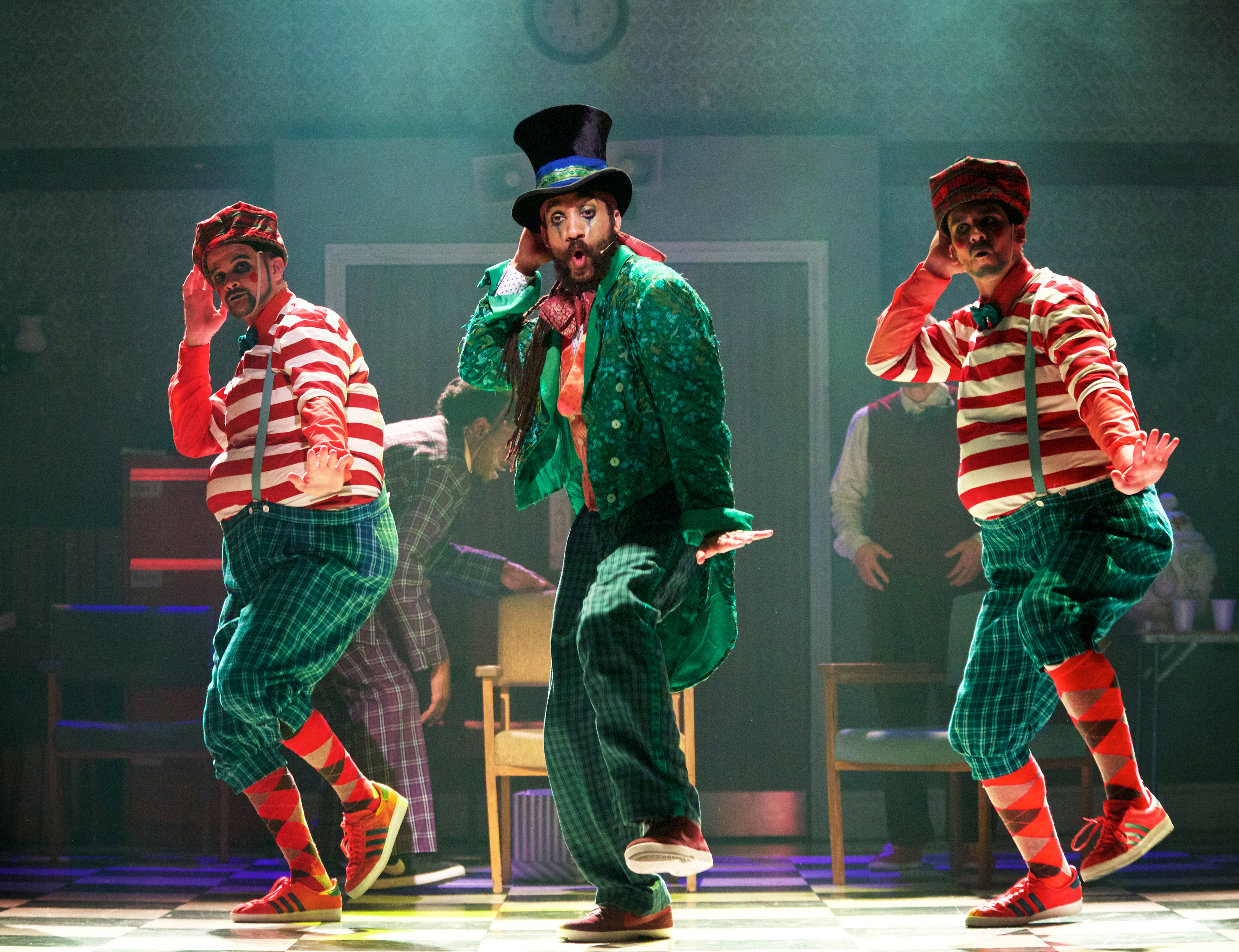 Three dancers are on stage: one in the centre is wearing a bright green coat and a top hat, two either side of him are wearing green trousers and a red and white striped top.