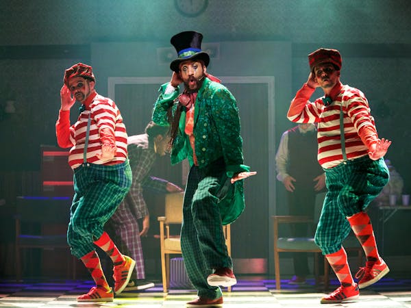 Three dancers are on stage: one in the centre is wearing a bright green coat and a top hat, two either side of him are wearing green trousers and a red and white striped top.