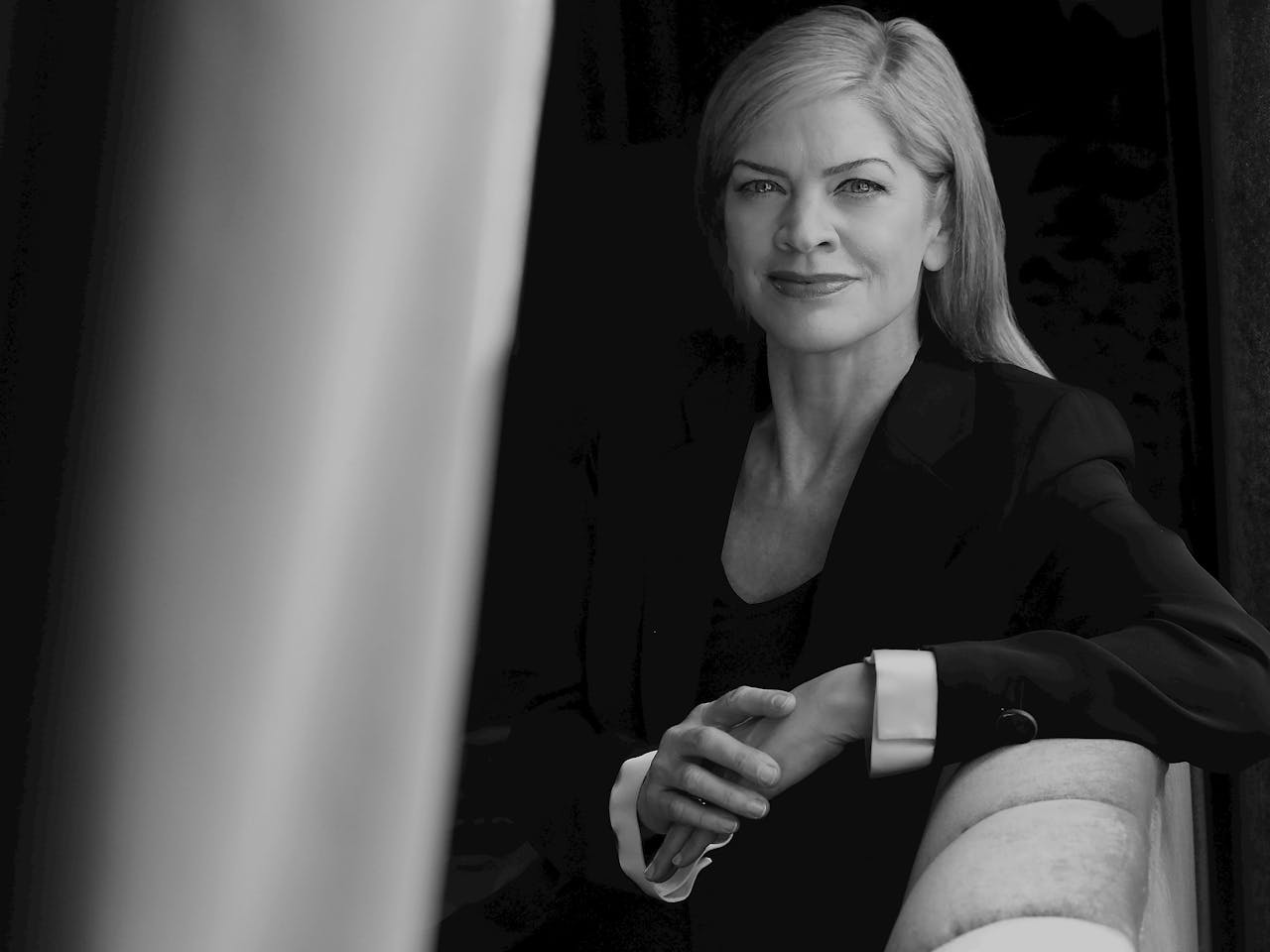 Black and white headshot of Canadian conductor Keri-Lynn Wilson. Keri has blonde air and is wearing a black suit jacket. She is sitting on a padded sofa and her arm is placed on the backrest as she smiles for the camera.