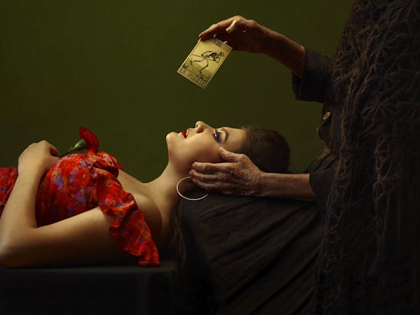 The opera singer Aigul Akhmetshina ilies down on her back wearing a red floral off the shoulder dress, purple trousers and large hooped earrings. Behind her stands a person wearing a black cloak who is only visible from the neck down. Their left hand is at the temple of the person lying down and their right hand holds up a tarot card with a skeleton on that reads la muerte signifying death.