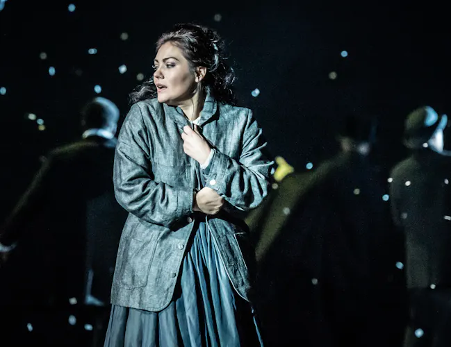 Ailyn Pérez as Mimì in La bohème at the Royal Opera House. Ailyn has long brown hair, and is wearing a costume with a grey jacket and dress. She is looking away to the side as she clasps her coat tightly as snow falls around her. (1 of 4)