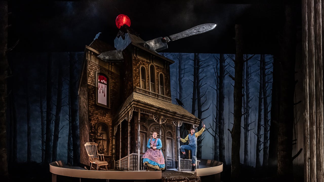 A looming house topped with a glowing cherry has a large knife cutting through the roof as if it was a cake. Two performers sing in bright costumes as the fairytale characters Hansel and Gretel in a production at the Royal Opera House. The header reads 2024/25 Accessible Performances.