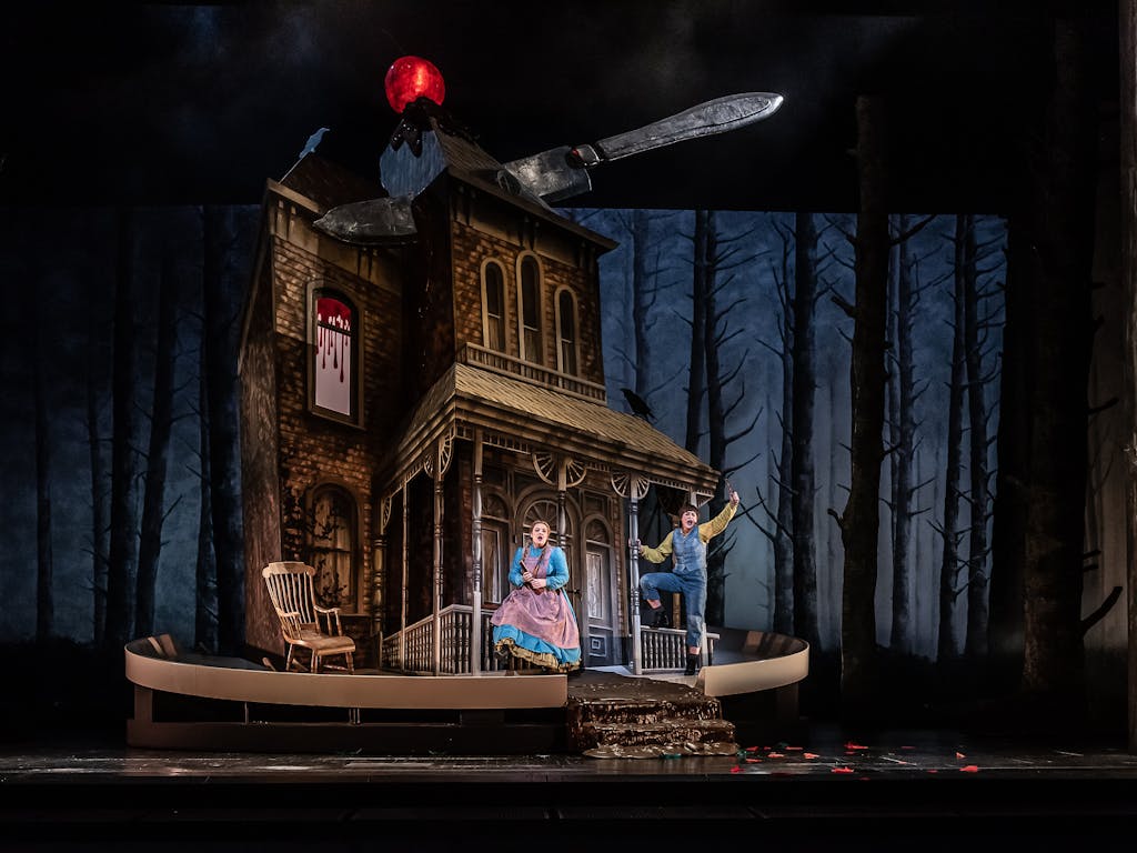 A looming house topped with a glowing cherry has a large knife cutting through the roof as if it was a cake. Two performers sing in bright costumes as the fairytale characters Hansel and Gretel in a production at the Royal Opera House.