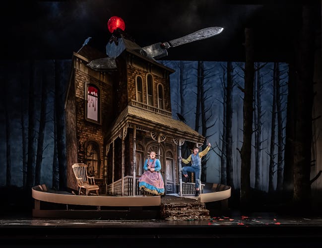 A looming house topped with a glowing cherry has a large knife cutting through the roof as if it was a cake. Two performers sing in bright costumes as the fairytale characters Hansel and Gretel in a production at the Royal Opera House.