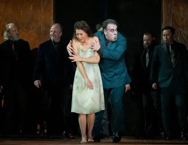 A woman in a short white dress stands on stage. A man in a a blue suit has his arms around her shoulders, gripping tightly. Men in dark suits stand in the shadows behind them.