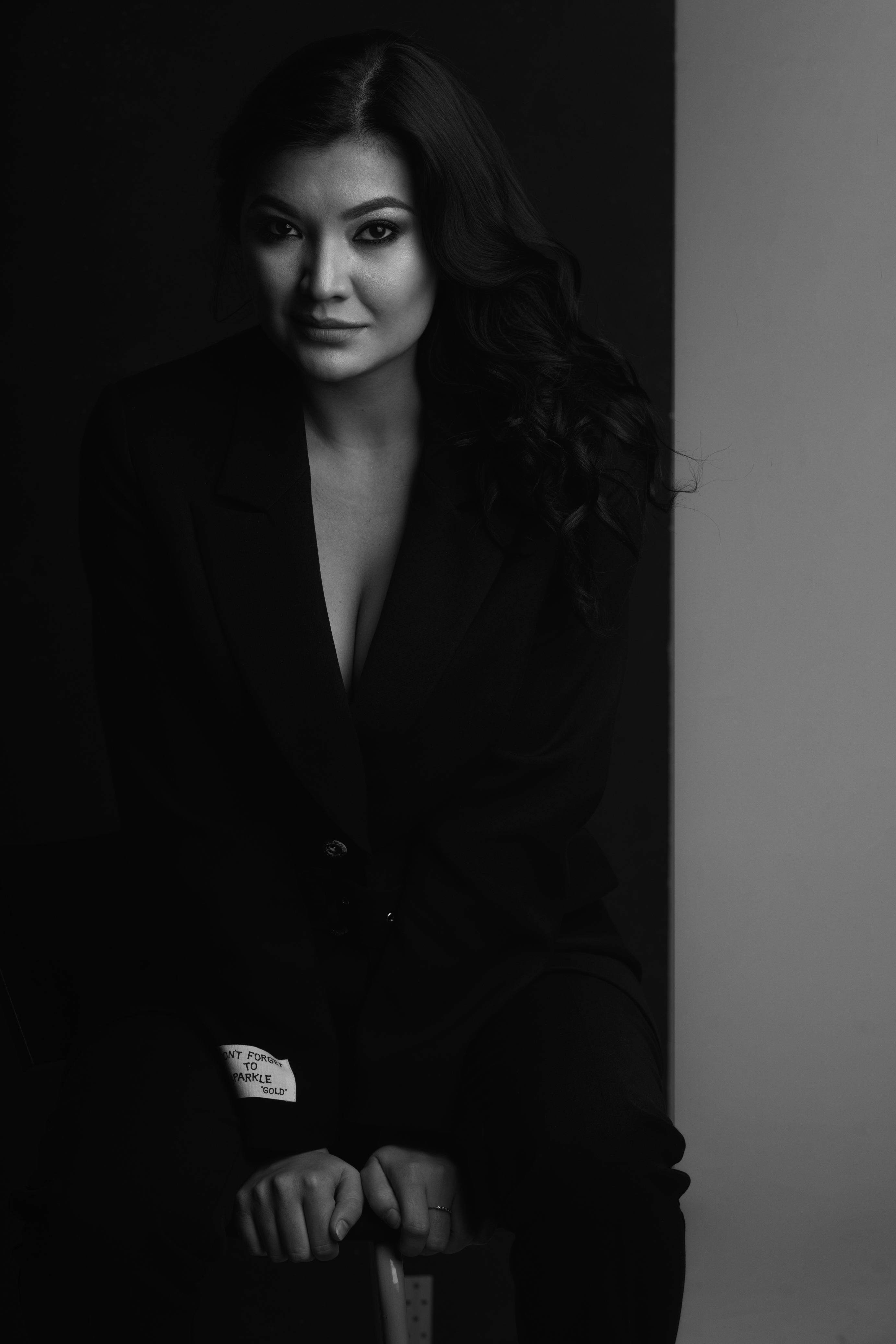 A black and white headshot of the opera singer Aigul Akhmetshina. She leans against a chair wearing a black top with her long dark hair worn down.