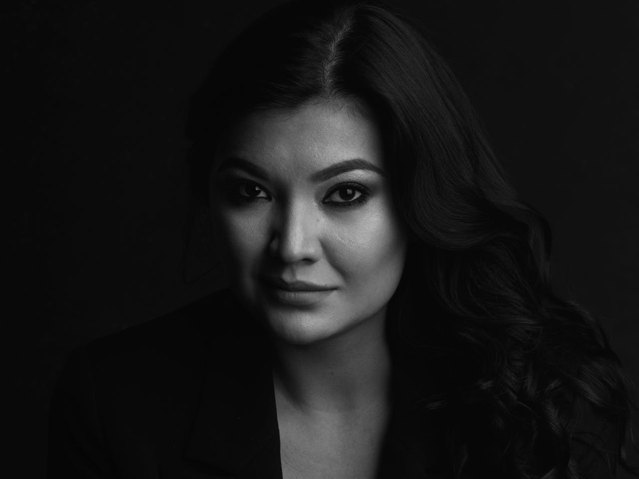 A black and white headshot of the opera singer Aigul Akhmetshina. She leans against a chair wearing a black top with her long dark hair worn down.
